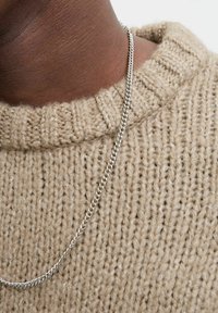 Silberne Ketten-Halskette, die auf einem strukturierten, beigen Strickpullover mit geripptem Kragen liegt. Die Materialien kontrastieren sanft in Farbe und Textur.