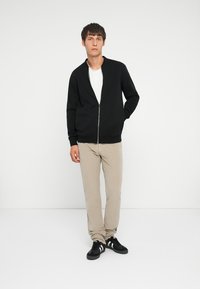 Black zip-up sweater with a textured fabric, white t-shirt underneath, beige pants, and black sneakers. Simple and modern design.