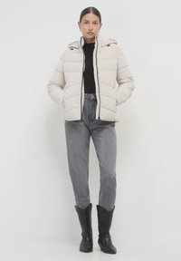 Eine Frau trägt eine beige Steppjacke, ein schwarzes Oberteil, graue High-Waist-Jeans und schwarze bestickte Cowboy-Stiefel und steht vor einem weißen Hintergrund.