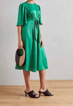 Kobieta w zielonej sukience midi z marszczonym paskiem, trzymająca okrągłą czarno-beżową torebkę, nosząca czarne sandały na obcasie, stojąca na drewnianej podłodze.