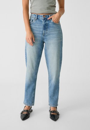 Vrouw draagt lichtblauwe jeans met hoge taille en een vervaagde voorkant, gecombineerd met zwarte puntige platte schoenen met gespen en studs als details.