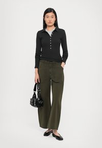 Zwarte langemouwen top met een kraag en knoopdetails, gecombineerd met olijfgroene wijde broek. Geaccessoireerd met een kleine zwarte tas.