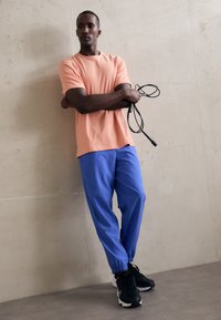 Peach short-sleeve t-shirt paired with blue tapered pants, held together with black athletic shoes, and a jump rope in hands, against a neutral wall.