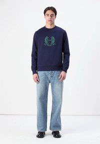 Navy blue sweatshirt with a crew neck and green logo design, paired with light blue straight-leg jeans and black shoes.