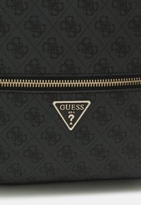 Tejido negro con un monograma texturizado repetido de "GS", cierre dorado y logo triangular de Guess USA con un signo de interrogación en la superficie de la bolsa.