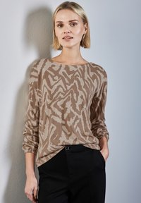 Beige Langarmshirt mit einem wellenförmigen abstrakten Muster in einem helleren Beige. Mit einem weiten Ausschnitt und gepufften Manschetten, kombiniert mit schwarzen Hosen.