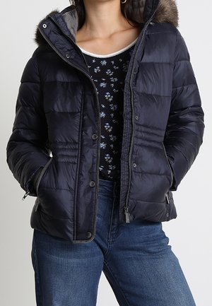 Kvinde iført mørkeblå quiltet jakke med pelsforet hætte over sort blomstret top og blå jeans, hænder i jakkelommer.