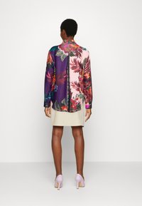 Camisa de manga larga con un vibrante patrón floral en seda morada y rosa. El diseño en la espalda tiene una costura central; combinada con una falda beige y tacones color lavanda.