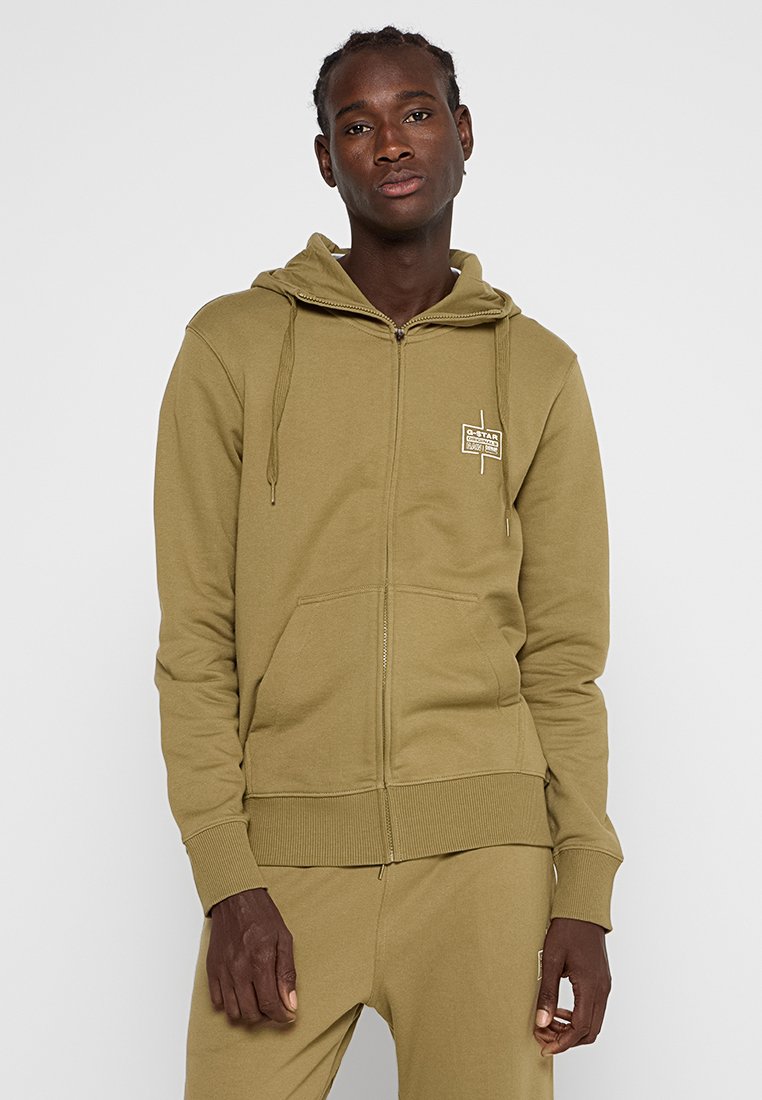 Olivgrüner Reißverschluss-Hoodie aus weichem Stoff, mit einer Kängurutasche, gerippten Bündchen und einem kleinen Logo-Patch auf der Brust.