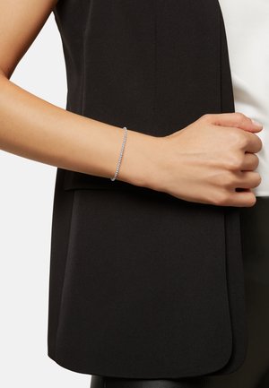 Pulsera de plata con piedras claras, diseño delgado, textura suave, llevada en una muñeca bronceada, combinada con una prenda negra.