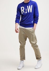 Blå sweatshirt med stora vita "R"- och "W"-bokstäver, i kombination med ljusbeiga cargobyxor och vita sneakers. Avslappnad passform, ribbade ärmslut.