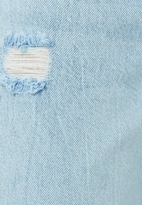 Tela de mezclilla azul claro con un desgarro deshilachado, mostrando hilos blancos debajo. La textura es suave, con un patrón tejido estándar.