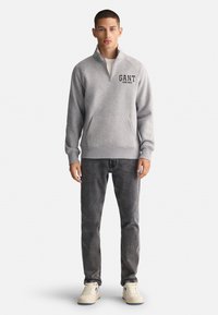 Szara bluza z ćwierć zamkiem, logo GANT na lewej piersi, w połączeniu z ciemnoszarymi dżinsami i beżowymi trampkami.