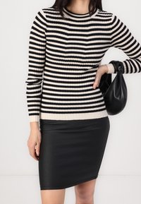 Femme portant un pull à manches longues rayé noir et blanc, jupe en cuir noir, tenant un sac à main en cuir noir à ses côtés.