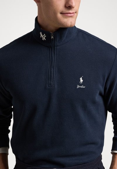 Άνδρας που φοράει μπλε σκούρο πουλόβερ με φερμουάρ μέχρι τον καρπό, με λογότυπο Polo Ralph Lauren και έμβλημα των New York Yankees στον γιακά.