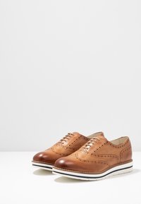 Brogues in pelle marrone con dettagli perforati, punta rotonda e suola bianca con striscia nera. Texture liscia, design classico a punta di ala.