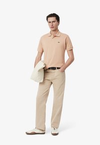 Polo en melocotón, de manga corta, con un logo verde. Combinado con pantalones beige claro y zapatillas blancas. Sosteniendo un bolso crema.