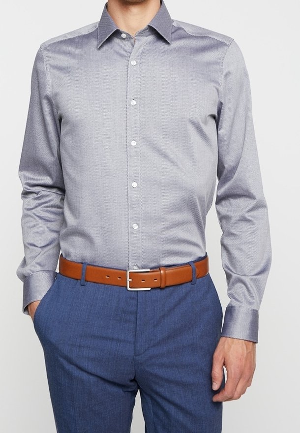 Homme portant une chemise à manches longues boutonnée de couleur gris clair avec motifs, rentrée dans un pantalon bleu avec une ceinture en cuir marron.