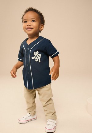 Maglietta da baseball blu navy con bordo bianco e decorativo lettering "LA", abbinata a pantaloni beige e sneakers bianche con cinturini in velcro.