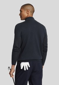 Navy pullover sweater, knit texture, medium-weight, featuring a high collar. White golf gloves tucked in back pocket, black golf club held in hand.