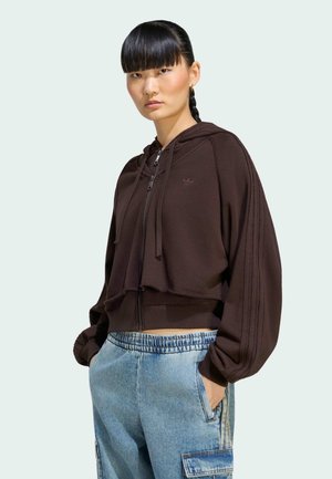 Giovane donna con i capelli intrecciati che indossa una felpa cropped marrone scuro e jeans cargo azzurri con le mani nelle tasche.