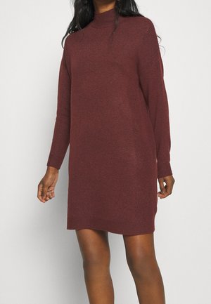 Kvinde iført en langærmet, knælang, ribstrikket vinrød sweaterkjole mod en ensfarvet lys baggrund.