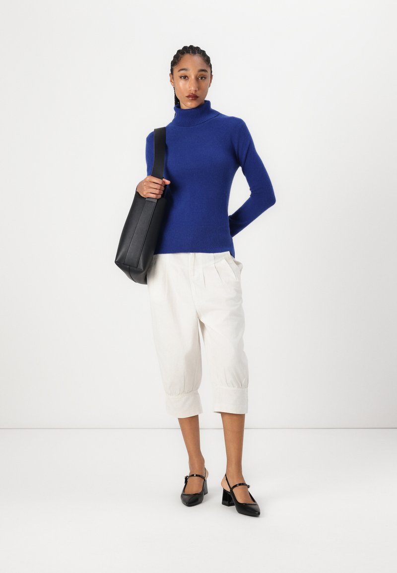 Un pull col roulé en tricot bleu, un pantalon cropped crème et des escarpins à talons ouverts noirs. Le mannequin porte un sac à épaule noir. Design minimaliste.