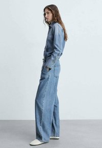 Ritja denirti vaaleansinisena, jossa on nappikiinnitys edessä, pitkät hihat ja leveät, löysät lahkeet. Kankaan pinta on teksturoitu ja siinä on näkyviä sauman yksityiskohtia.