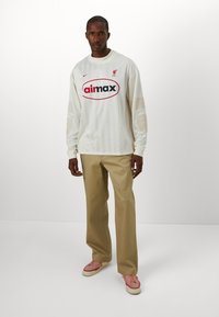 Maglietta a maniche lunghe color crema in jersey con logo rosso e grafica. Notevole motivo a righe e design del testo. Abbinata a pantaloni beige chiaro e sneakers rosa.