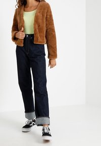 Giacca in pelliccia sintetica marrone sopra una canotta verde chiaro, abbinata a jeans a vita alta blu scuro e sneakers nere con suola bianca.