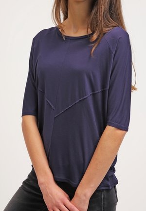 Granatowa bluzka z krótkim rękawem z ukośnym szwem; wykonana z miękkiego, lekkiego materiału; ma okrągły dekolt i luźny krój.