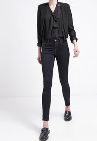 Blouse noire avec un nœud papillon et des manches courtes, associée à un jean skinny en denim foncé et des chaussures noires à enfiler, créant un look ajusté.