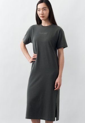 Jeune femme aux longs cheveux noirs portant une robe gris charbon, à manches courtes, longueur genou, avec un texte subtil « Barbour International ».