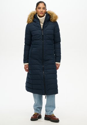 Kobieta ubrana w długi granatowy pikowany płaszcz z kapturem wykończonym futrem, jasnoniebieskie dżinsy, brązowe buty i kremowy sweter z golfem.