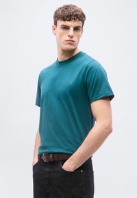 Νεαρός άνδρας που φοράει πετρόλ κοντομάνικο μπλουζάκι και σκούρο τζιν με καφέ ζώνη, στέκεται με τα χέρια στις τσέπες μπροστά σε απλό φόντο.