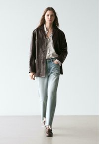 Braune Lederjacke über einem weißen, gestreiften Hemd, kombiniert mit hellblauen Jeans und braunen Schuhen. Die Jeans haben ausgefranste Säume.