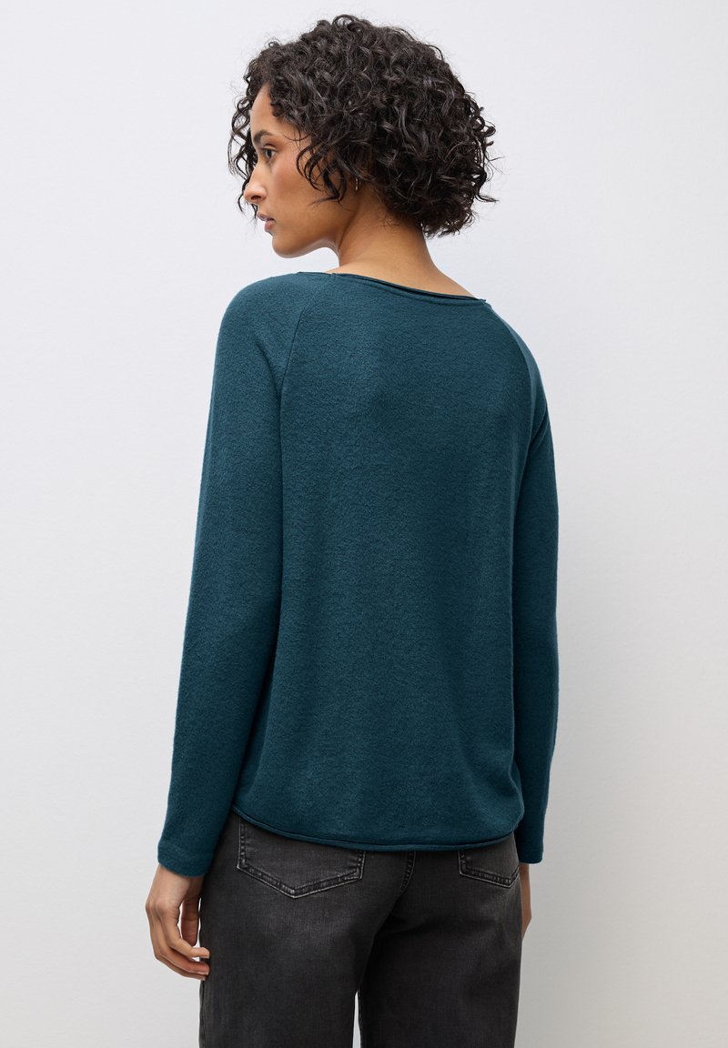 Türkiser langärmeliger Pullover mit Rundhalsausschnitt und Raglanärmeln. Weiche Textur und entspannte Passform. Getragen mit dunkelgrauen Jeans.