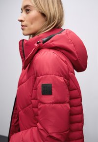 Chaqueta de plumas roja con diseño acolchado, con un cuello alto y un parche de logo negro en el hombro, que muestra un material liso y brillante.