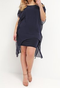 Robe bleu marine en voile transparent avec un ourlet incurvé, design sans manches et couche intérieure ajustée. Associée à des talons nus à lanières.