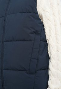 Námornícky modrá prešívaná bunda s prednou kapsou vedľa blízkeho detailu rukáva smotanovo bieleho pleteného svetra.