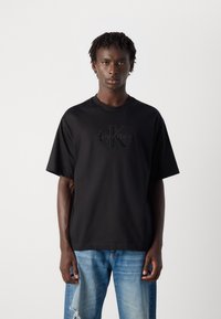 Crna majica kratkih rukava s okruglim izrezom, s utisnutim logotipom Calvin Klein na prednjoj strani. Uklopljena s svijetloplavim istrošenim trapericama.
