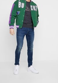 Zelená bomber bunda s fialovými detaily a nápisem "BUCKS", spárovaná s tmavě šedým tričkem, modrými džíny a bílými teniskami.