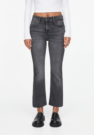 Woman wearing high-waist faded black cropped flare jeans, white long-sleeve top, and black slip-on shoes, standing against white background.