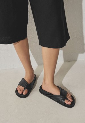 Ben iført sorte korte bukser og sorte krydsede slidesandaler, stående på en lys gulv mod en hvid væg.