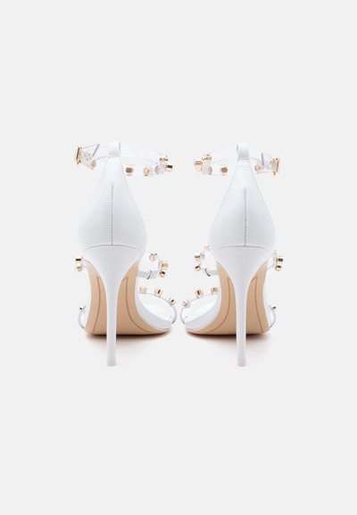 Λευκά πέδιλα με ψηλό τακούνι και διάφανους ιμάντες διακοσμημένους με χρυσά τρουκς, φωτισμένα από πίσω σε απλό ανοιχτό φόντο.