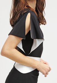 Svart klänning med vita accenter, med fjäderärmar och en skärning i midjan. Tyget är slätt och sitter tätt mot kroppen.