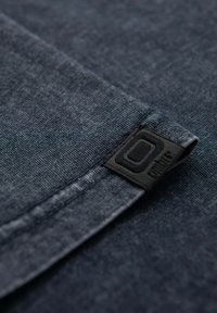 Tmavě modrý textil s texturovaným povrchem, obsahující černou tkanou cedulku s bílým logem. Látka má jemné variace v odstínu.