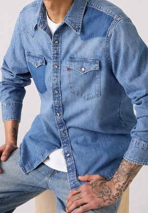 Mand iført lyseblå denimskjorte med hvide knapper og brystlommer, siddende; tatoveringer synlige på underarme og hænder.