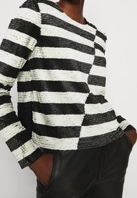 Personne portant un haut à manches longues rayé horizontalement noir et blanc avec des sequins texturés et un pantalon noir, les mains dans les poches.