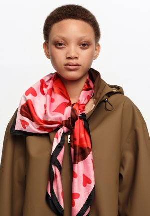 Joven con cabello corto y rizado, vistiendo una chaqueta marrón con cremallera y una bufanda rosa con diseño de corazones rojos atada al cuello.